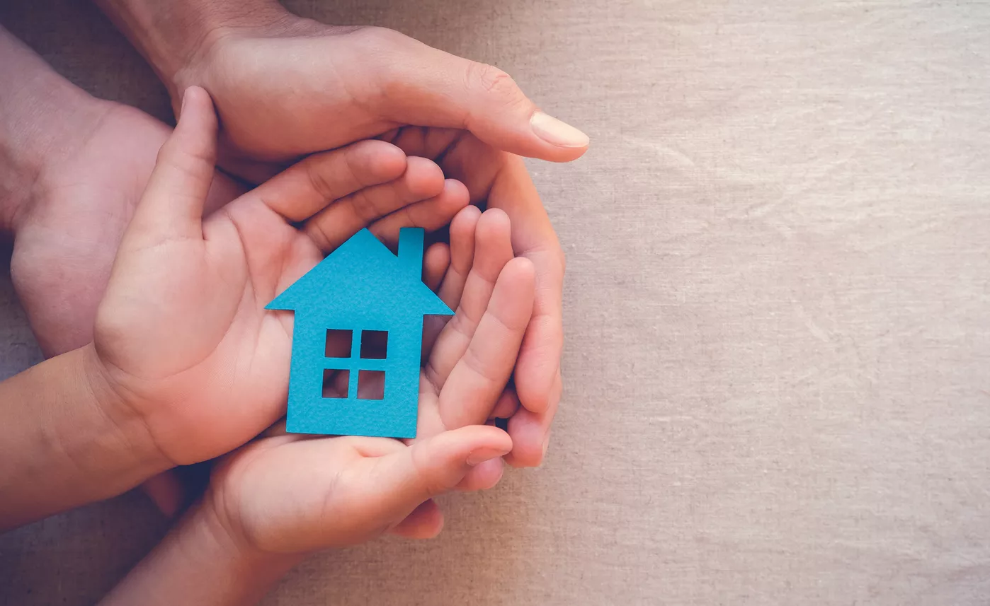  A larger pair of hands cradles a smaller pair of hands, which are cradling a cutout of a house.
