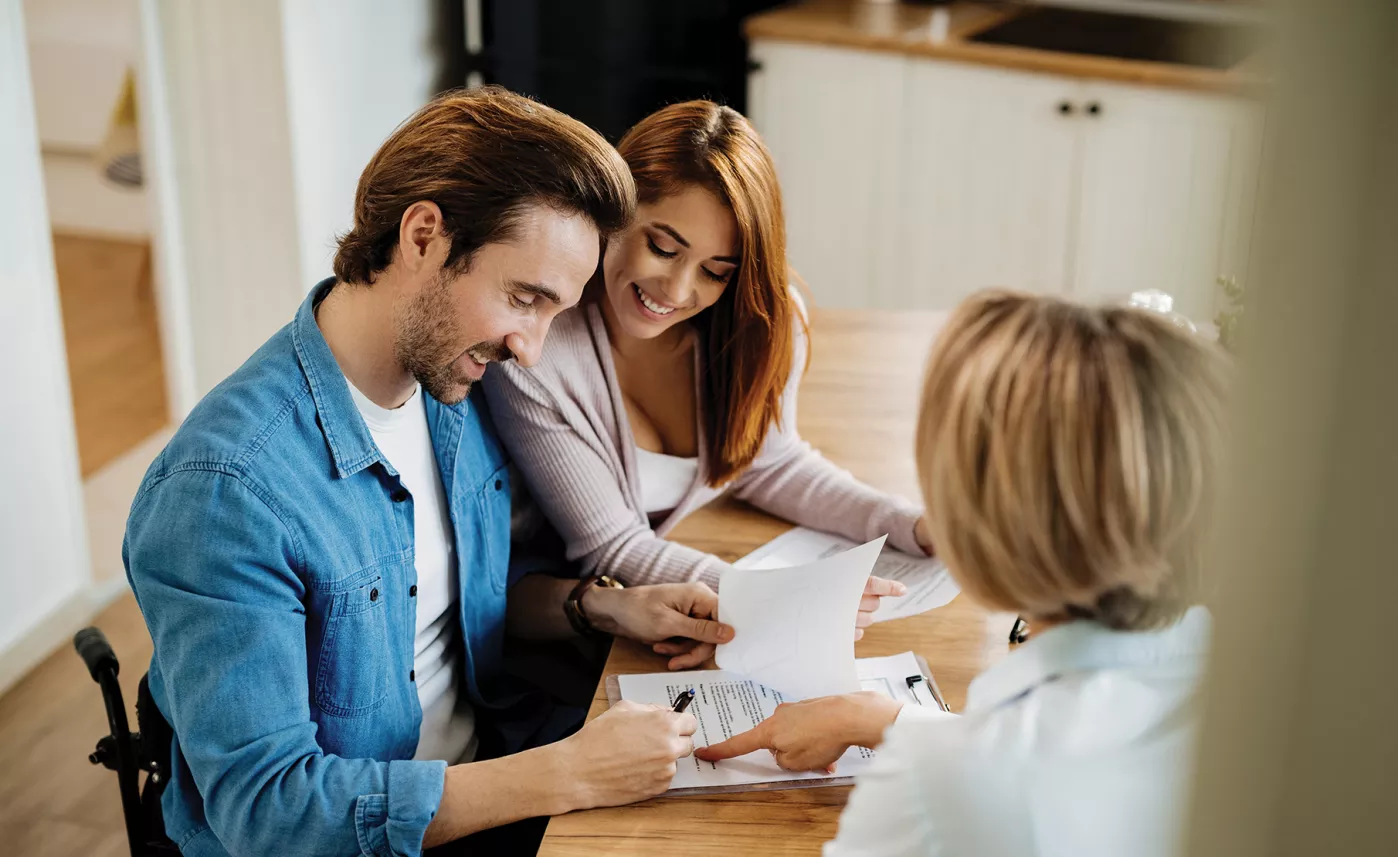  A couple sitting at their kitchen table signing paperwork with their financial advisor.
