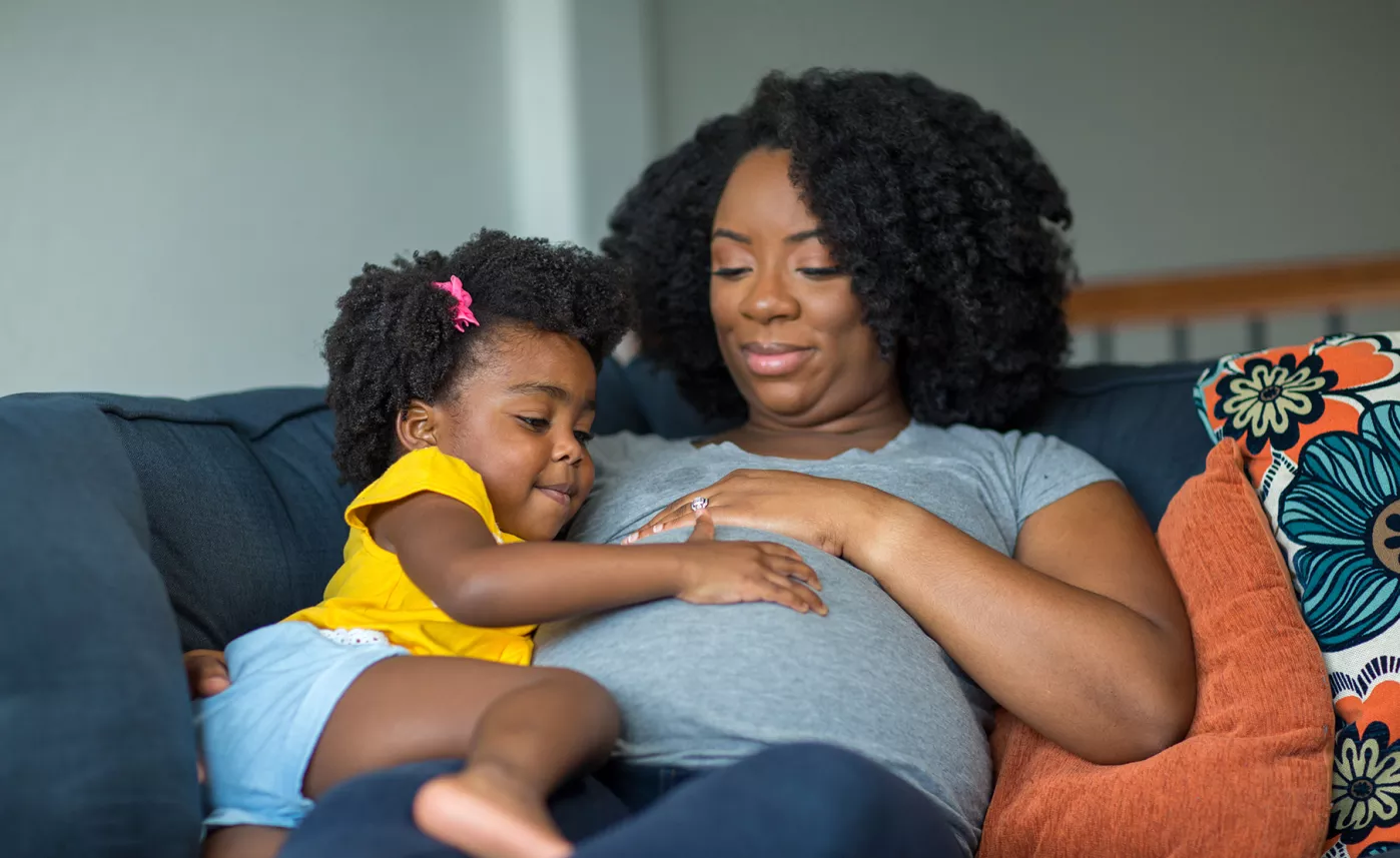  Une mère enceinte est étendue sur le canapé avec sa fille, la main de chacune posée sur le ventre de la mère.
