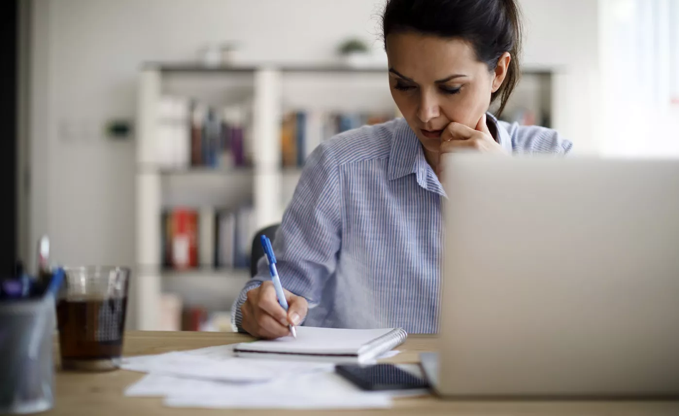  A woman writes notes in a notebook in front of her laptop, carefully calculating her financial strategy.
