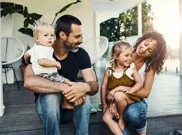  Une jeune famille (le père, la mère et les deux jeunes enfants) est assise sous la véranda et rie.
