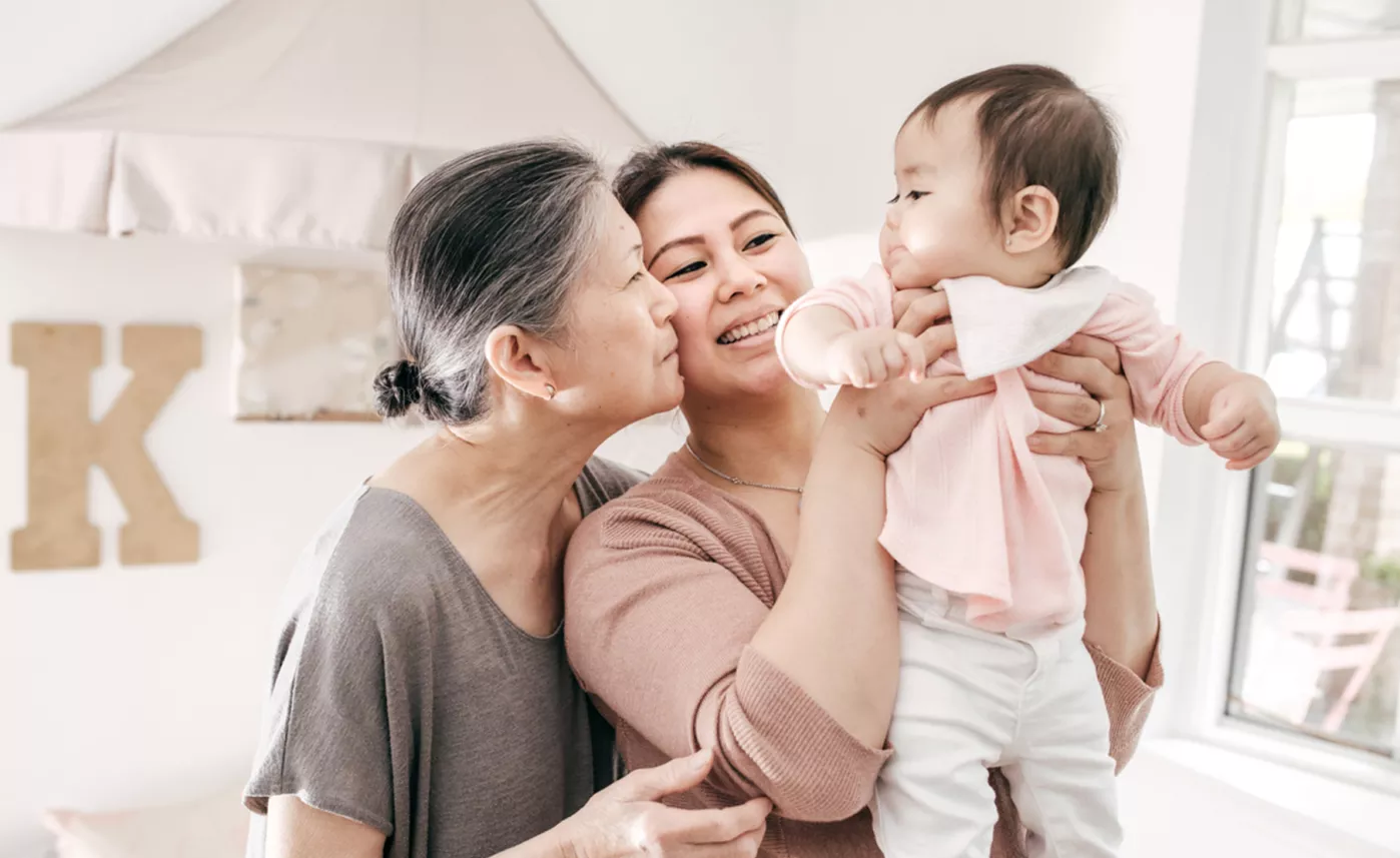  Mother smiling, holding toddler child, while grandmother looks on smiling
