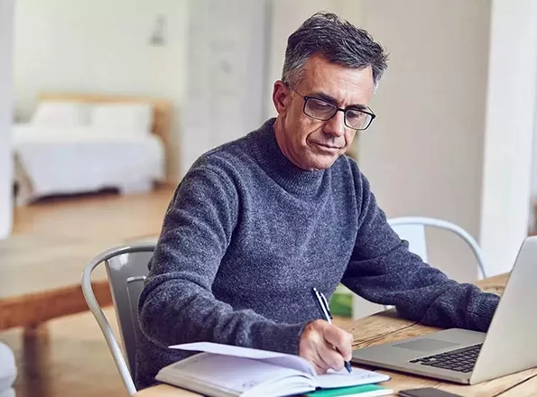  A man checks his finances in front of his laptop at his kitchen table, and takes notes in a logbook.
