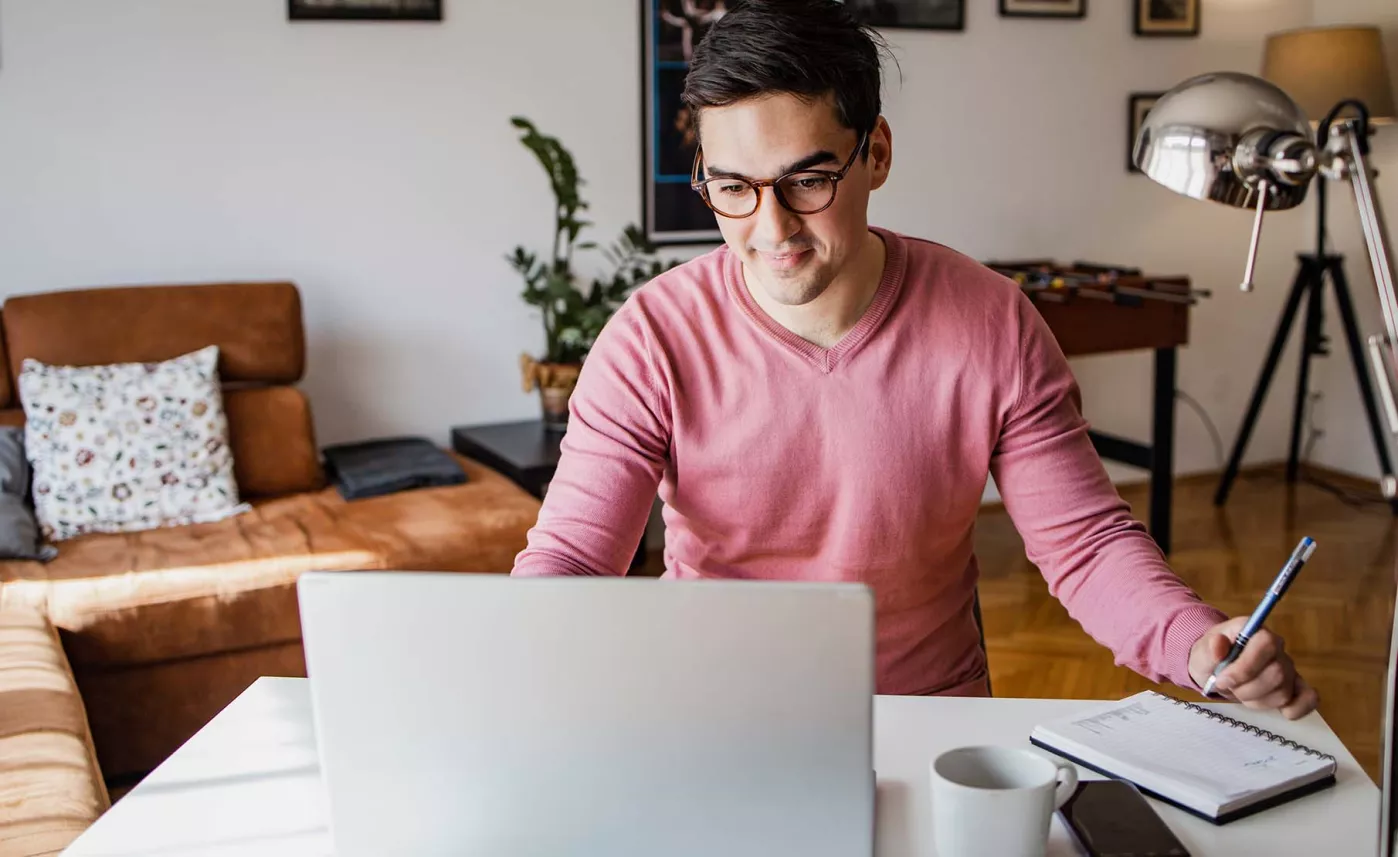  A person wearing glasses look at their laptop and writing in a notebook.
