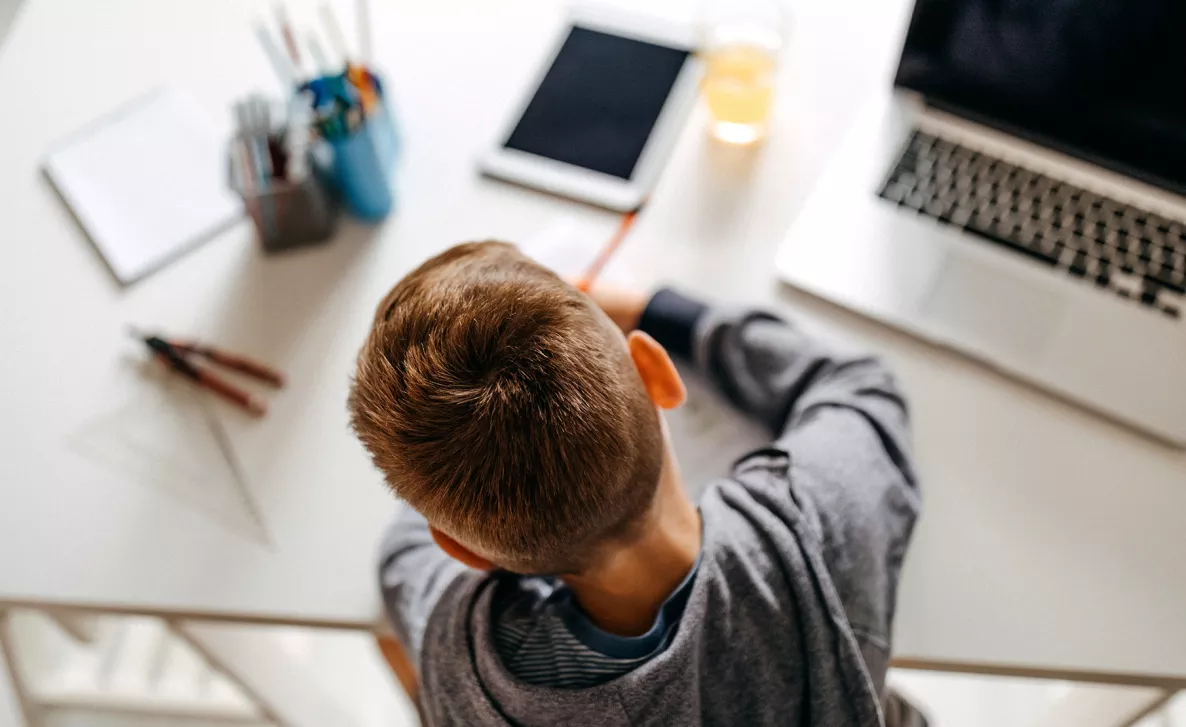  Un jeune garçon fait ses devoirs à son bureau, son ordinateur portable ouvert devant lui.
