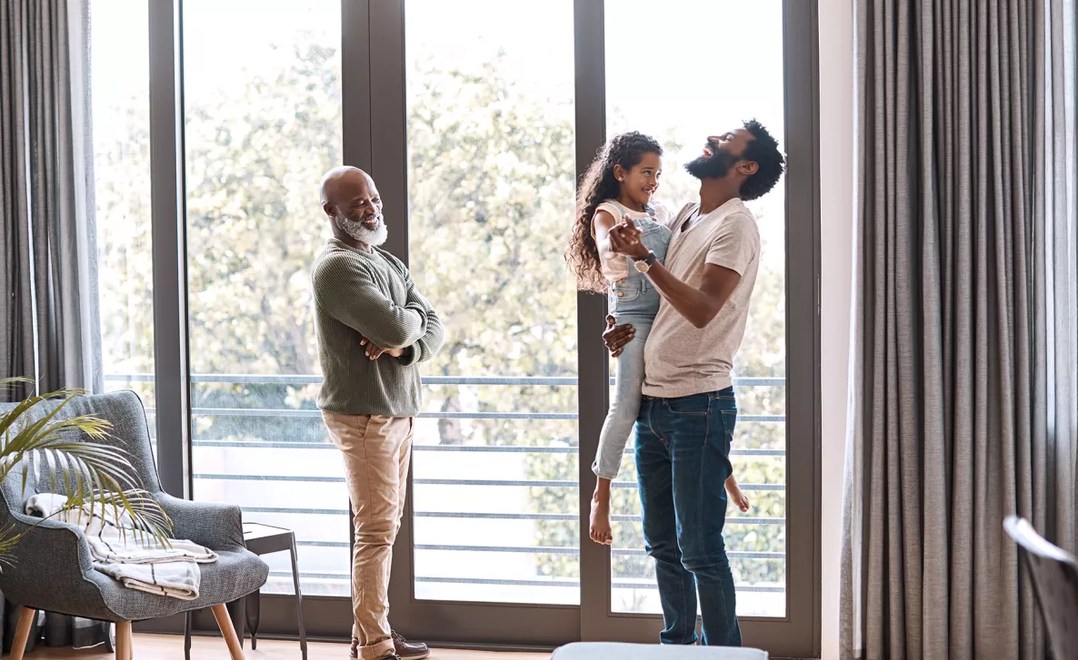  A young father hold his young daughter in a sunny living room, while a grandfather smiles at them.
