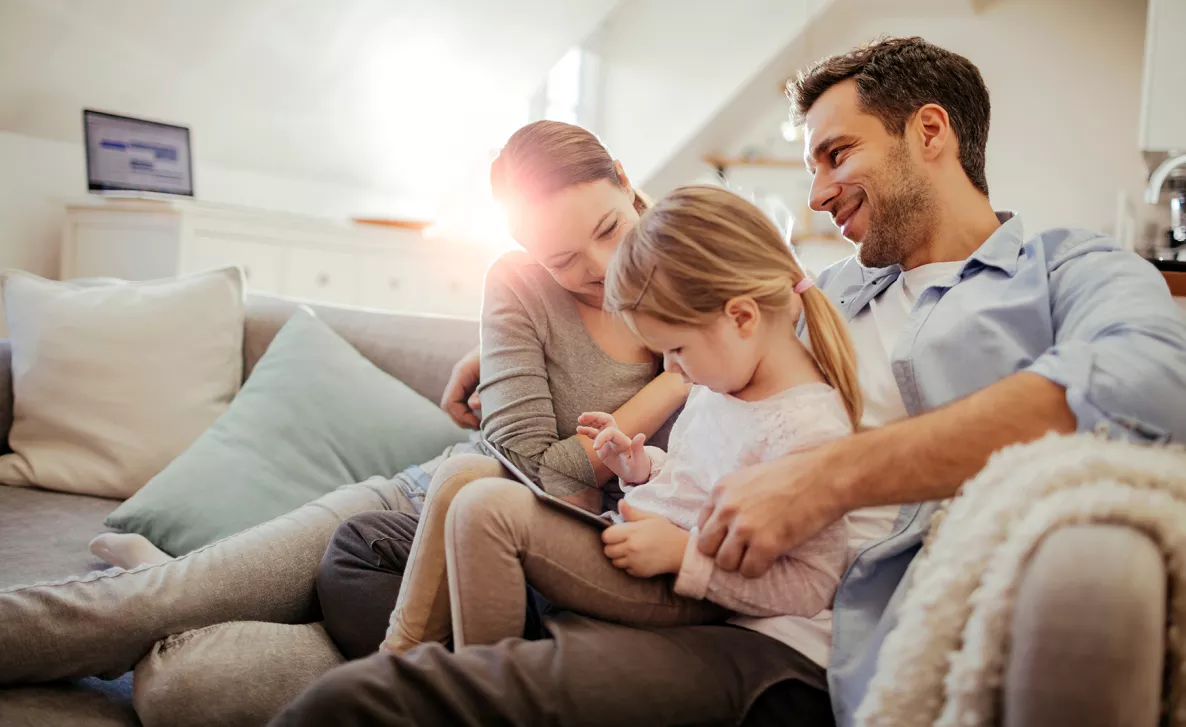  Young parents sit with their toddler daughter as she plays on her tablet.
