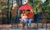 A young family sits under an umbrella on a bench outdoors.