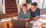 A young couple reviews financial information on their laptop, with notebooks spread out in front of them.