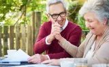 An elderly couple smile and hold hands as they look at documents on their porch.