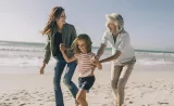 A retired woman laughs on the beach with her adult daughter and young granddaughter.