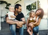 Une jeune famille (le père, la mère et les deux jeunes enfants) est assise sous la véranda et rie.