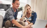 A young couple checks their financial account on their laptop with a puppy on their laps.