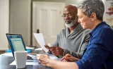 Un couple en âge de prendre sa retraite examine des documents sur leur ordinateur portable dans leur cuisine.